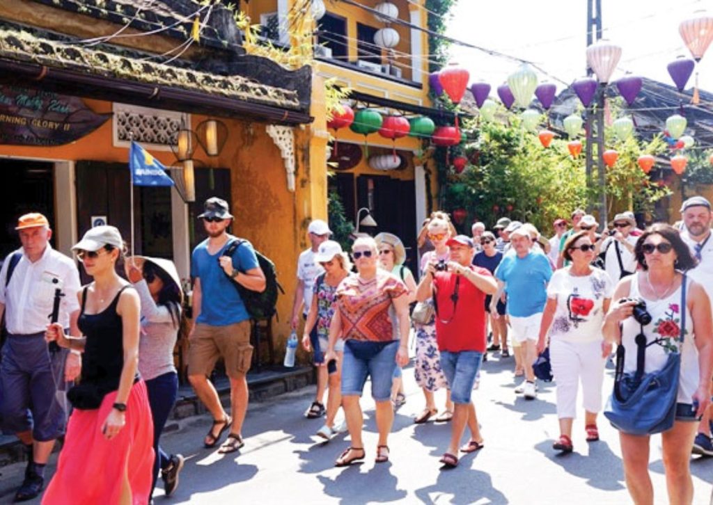 Crowds of tourists exploring the ancient town of Hoi An under colorful lanterns on a bright, sunny day